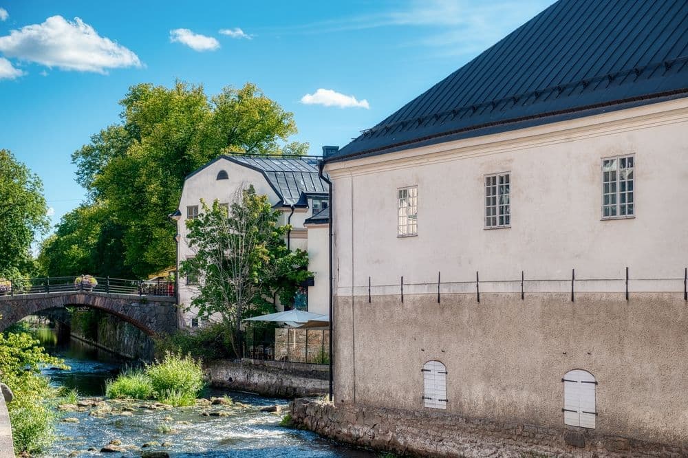 Vitputsade byggnader vid ett vattendrag med stenbro och grönska, fotograferat i dagsljus i Uppsala.
